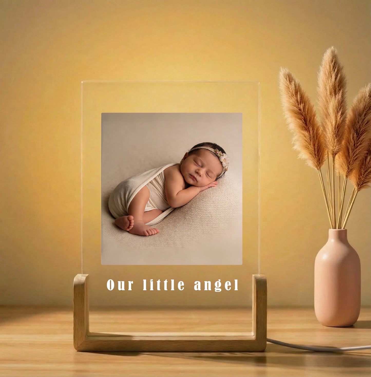 Framed photo of a baby with 'Our little angel' text on a wooden stand, next to a vase with dried plants.